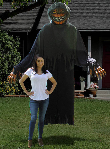 Eine Frau steht vor einer großen, auffälligen Halloween-Dekoration auf einem Rasen – einer grinsenden Gestalt mit Kapuze, die genau wie das „Horror-Kürbis-Kostüm oder -Dekoration, ca. 240 cm“ aussieht.