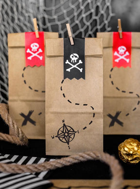 Im Set sind sechs braune Papier-„Geschenktüten Piratenparty“ enthalten, die jeweils mit Piratenmotiven wie Totenkopf-Aufklebern und Schatzkarten-Motiven verziert sind. Im Vordergrund sind Seile und goldverpackte Schokoladenmünzen zu sehen – diese Tüten eignen sich ideal als umweltfreundliche Geschenktüten für eine aufregende Piratenparty zum Kindergeburtstag.