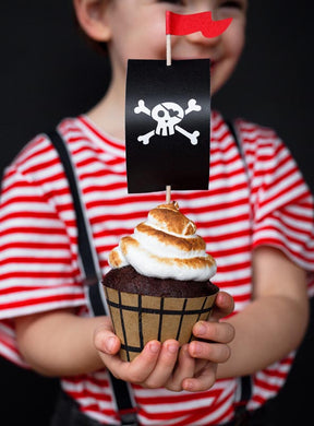Ein Kind in einem rot-weiß gestreiften Shirt hält einen Cupcake in der Hand, der mit einer Piratenflagge und Zuckergussstreifen verziert ist. Der Cupcake befindet sich in festlichen Backförmchen aus dem „Cupcake Deko Pirat 6er-Set Piratenschiff“ – ideal für Cupcakes zum Thema Piraten – und steht vor einem dunklen Hintergrund.