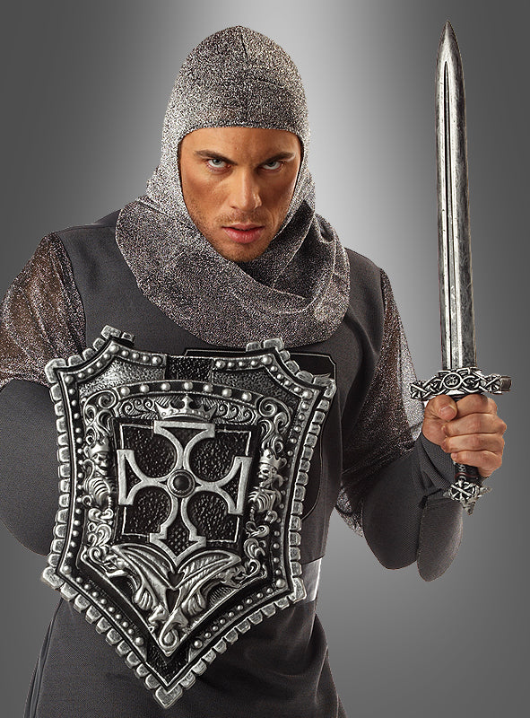Ein als mittelalterlicher Ritter gekleideter Mann hält das „Kreuzritter-Schwert 60 cm mit Schild“ – bekleidet mit Kettenhemd und mit ernstem Gesichtsausdruck – vor einem grauen Hintergrund.