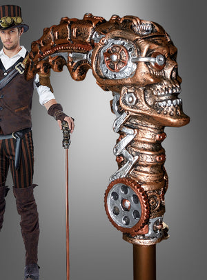 Ein Mann in einem Steampunk-Kostüm hält den „Gehstock Steampunk Alien“ in der Hand, einen Spazierstock mit einem metallischen, von Zahnrädern verzierten Griff in Form eines mechanischen Schädels – das perfekte Spazierstock-Accessoire, um seinen unverwechselbaren Look zu vervollständigen.