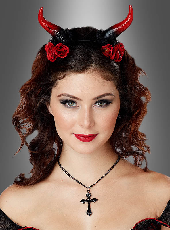 Eine Frau mit welligem braunem Haar trägt rote Teufelshörner mit Rosen, eine schwarze Kreuzkette und dunkles Make-up und posiert vor einem grauen Hintergrund mit Farbverlauf.