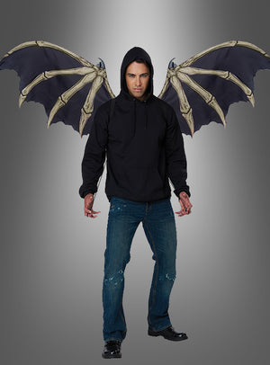 Vor einem schlichten Hintergrund mit Farbverlauf steht ein Mann in einem schwarzen Kapuzenpulli und blauen Jeans, an dessen Rücken große Dämonen-Skelettflügel befestigt sind – ein auffälliges Halloween-Cosplay.