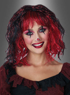 Vor einem grauen Farbverlaufshintergrund lächelt eine Frau in die Kamera und präsentiert dabei ein auffälliges rot-schwarzes Clown-Make-up sowie eine rote Nase. Dazu trägt sie die Perücke „Pilotta“ mit rot-schwarzen Lametta-Strähnen – ein farbenfrohes Karnevalsaccessoire.