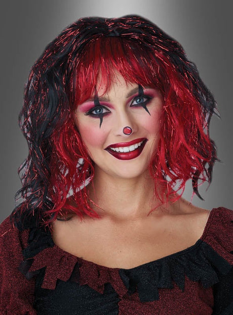 Vor einem grauen Farbverlaufshintergrund lächelt eine Frau in die Kamera und präsentiert dabei ein auffälliges rot-schwarzes Clown-Make-up sowie eine rote Nase. Dazu trägt sie die Perücke „Pilotta“ mit rot-schwarzen Lametta-Strähnen – ein farbenfrohes Karnevalsaccessoire.