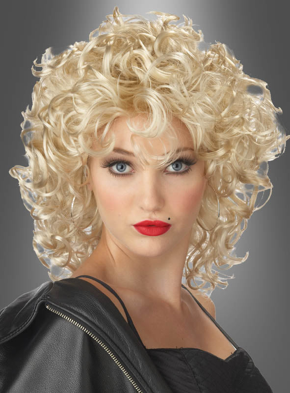 Eine Frau mit auffälligem Make-up, rotem Lippenstift und einem Schönheitsfleck, ganz in Schwarz gekleidet und vor einem grauen Hintergrund mit Farbverlauf posierend. Sie rundet ihren markanten Look mit der blonden „Bad Girl Sandy“-Perücke ab, die ihr das unverwechselbare Flair eines echten Lockenkopfs verleiht.