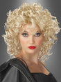Eine Frau mit auffälligem Make-up, rotem Lippenstift und einem Schönheitsfleck, ganz in Schwarz gekleidet und vor einem grauen Hintergrund mit Farbverlauf posierend. Sie rundet ihren markanten Look mit der blonden „Bad Girl Sandy“-Perücke ab, die ihr das unverwechselbare Flair eines echten Lockenkopfs verleiht.