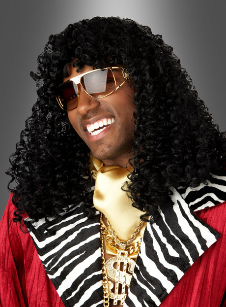Eine lächelnde Person mit langen, lockigen schwarzen Haaren, die zu einer klassischen „Pimp Big Daddy“-Perücke frisiert sind, mit Sonnenbrille, einem rot-zebra-gestreiften Mantel, einem goldenen Schal und mehreren goldenen Halsketten, die vor einem einfarbigen Hintergrund mit Farbverlauf posiert.