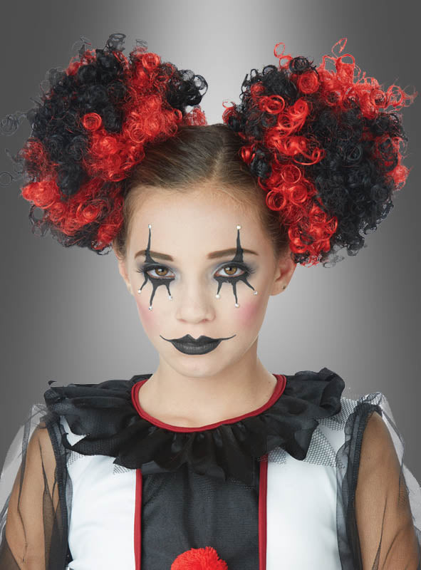 Ein junges Mädchen in einem Clownskostüm trägt eine auffällige schwarz-rote Lockenfrisur, die mit „Lockige Haarteile für Clownskostüm“ gestaltet wurde, sowie passende Pompons. Abgerundet wird ihr Look durch dunkles Augen-Make-up, schwarzen Lippenstift und einen Rüschenkragen, während sie vor einem grauen Hintergrund direkt in die Kamera blickt.