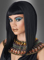 Eine Person in einem Kleopatra-Kostüm, das von einer altägyptischen Königin inspiriert ist, mit der „Schwarzen Kleopatra-Perücke“, auffälligem Augen-Make-up, goldenem Lippenstift und einem mit Edelsteinen besetzten Kragen, vor grauem Hintergrund.