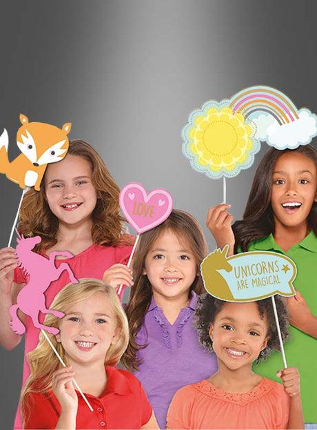 Fünf lächelnde Kinder halten vor einem grauen Hintergrund bunte Papierrequisiten aus dem „Fotoset Requisiten für Kinder“ in den Händen, darunter ein Einhorn, einen Fuchs, ein Herz, eine Sonne mit Wolke und ein Schild mit der Aufschrift „Einhörner sind magisch“ – perfekt als Partyzubehör für jede fröhliche Feier.