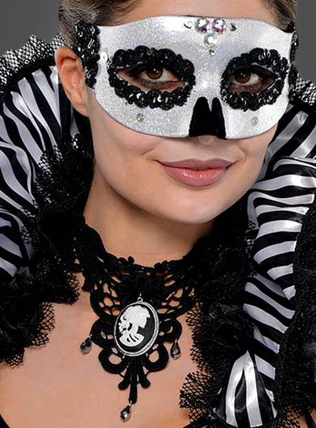 Eine Person, die eine schwarz-weiße Maskenballmaske, ein gotisches Halsschmuck-Cameo mit Skelettgesicht sowie ein schwarzes Outfit mit einem gestreiften Rüschenkragen trägt, vor einem dunklen Hintergrund.