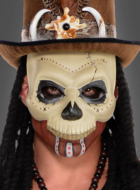 Eine Person, die die „Voodoo Maske Totenkopf für Damen und Herren“ trägt, mit aufgemalten Zähnen und Nähten, einem hohen Hut, der mit Hörnern und Federn verziert ist, Zöpfen und einer Perlenkette, vor einem schlichten grauen Hintergrund.