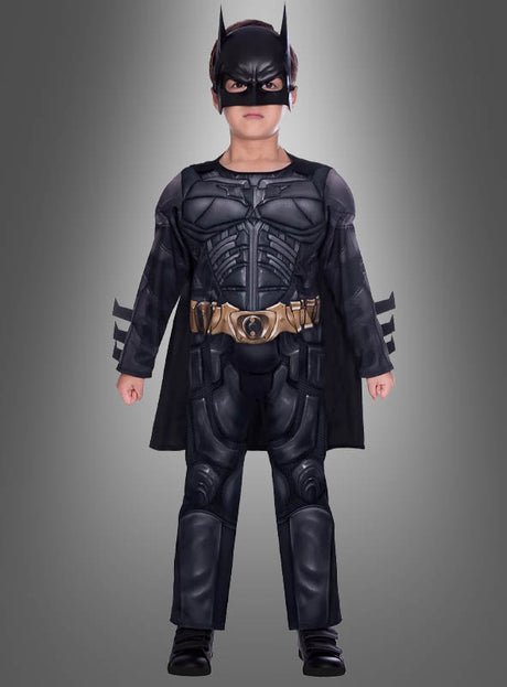 Ein Kind, das das „Batman Dark Knight“-Kostüm für Kinder trägt – mit Maske, Umhang und Werkzeuggürtel –, steht vor einem grauen Hintergrund.