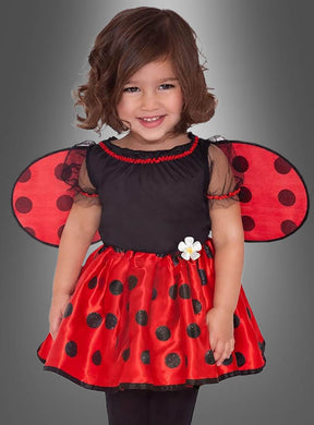 Ein kleines Kind trägt das Kinderkostüm „Kleiner Marienkäfer“ mit rot-schwarz gepunkteten Flügeln und einem passenden Rock und lächelt vor einem grauen Hintergrund. Dieses entzückende Kostüm ist perfekt für jedes kleine Mädchen, das sich gerne im bezaubernden Marienkäfer-Stil verkleidet.