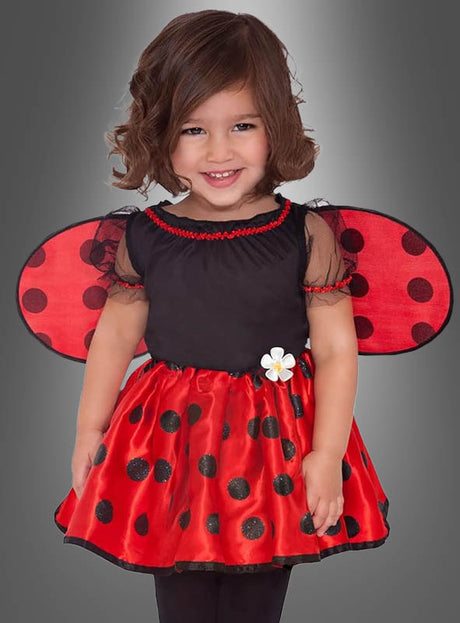 Ein kleines Kind trägt das Kinderkostüm „Kleiner Marienkäfer“ mit rot-schwarz gepunkteten Flügeln und einem passenden Rock und lächelt vor einem grauen Hintergrund. Dieses entzückende Kostüm ist perfekt für jedes kleine Mädchen, das sich gerne im bezaubernden Marienkäfer-Stil verkleidet.