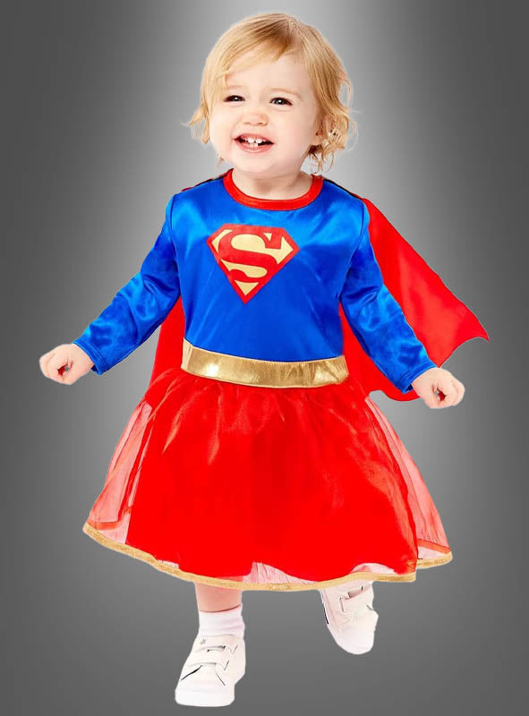 Ein kleines Kind, das das Baby-Kostüm „Supergirl-Kleid und Umhang“ trägt – ein blau-rotes Design mit einem S-Emblem und einem roten Umhang –, steht vor einem grauen Hintergrund in einem bezaubernden Superheldinnen-Outfit, das von der offiziellen DC Comics-Lizenz inspiriert ist.