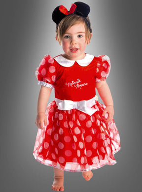 Ein kleines Kind steht barfuß da und trägt ein bezauberndes Minnie-Maus-Kleid aus rotem Stoff mit weißen Punkten sowie ein passendes schwarzes Stirnband mit Mausohren, das mit einer roten Schleife verziert ist. Im Hintergrund ist ein grauer Farbverlauf zu sehen.