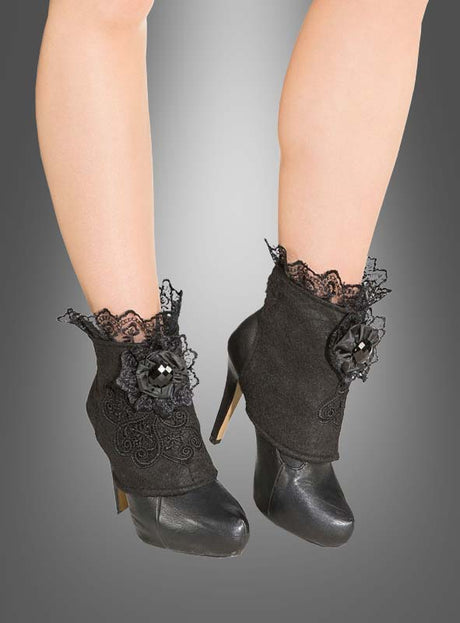 Vor einem grauen Hintergrund präsentiert das Model mit nackten Beinen schwarze Stöckelschuhe, die mit gotischen Schuhstulpen mit Spitze schwarz verziert sind – elegante schwarze Schuhstulpen mit Spitze und floralen Verzierungen.