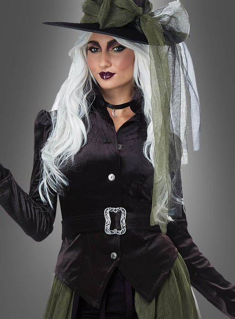 Eine Person, die eine schwarze Samtjacke, einen breitkrempigen Hut mit grünen und weißen Bändern sowie ein Halsband trägt, mit langen weißen Haaren und auffälligem Make-up, posiert vor einem grauen Hintergrund – ein bezauberndes, vielseitiges Hexenkostüm „Layla“, perfekt für jedes Hexenparty-Outfit oder als elegantes Karnevalskostüm.