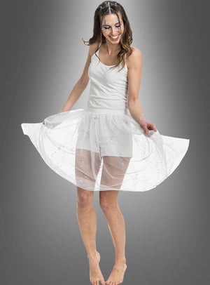 Eine Frau in einem weißen Kleid mit transparentem Rock lächelt und posiert barfuß vor einem grauen Hintergrund mit Farbverlauf, während sie ihren „Festival Tüllrock mit Glitzer“ hochhält.