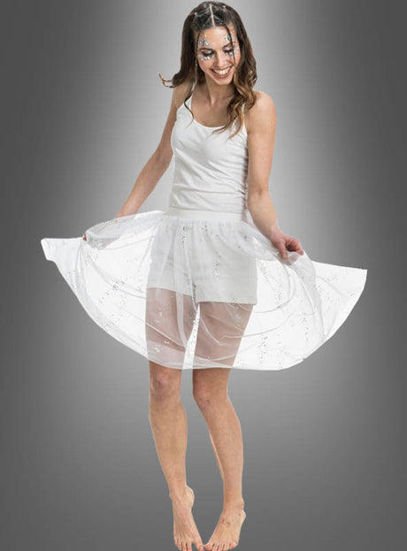 Eine Frau in einem weißen Kleid mit transparentem Rock lächelt und posiert barfuß vor einem grauen Hintergrund mit Farbverlauf, während sie ihren „Festival Tüllrock mit Glitzer“ hochhält.