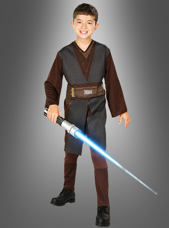 Ein lächelndes Kind in einem Anakin-Skywalker-Kostüm für Kinder hält ein blaues Lichtschwert in der Hand und steht vor einem schlichten grauen Hintergrund.