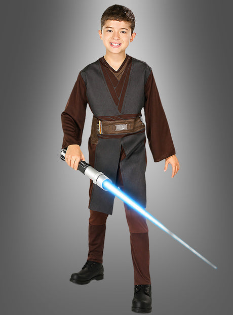 Ein lächelndes Kind in einem Anakin-Skywalker-Kostüm für Kinder hält ein blaues Lichtschwert in der Hand und steht vor einem schlichten grauen Hintergrund.