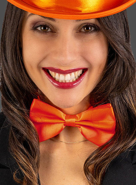 Eine Frau mit langen, dunklen Haaren trägt ein leuchtend orangefarbenes Satin-Fliege-Faschingsaccessoire als Fliege, dazu einen passenden Satin-Hut und ein schwarzes Outfit. Sie lächelt und steht vor einem schlichten Hintergrund.