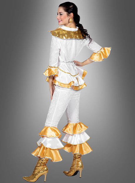 Eine Frau trägt das „Dancing Queen“-Disco-Kostüm (Zweiteiler) mit weißen und goldenen Rüschenärmeln und einer Hose mit Schlag, dazu passende goldene Stiefel mit Pailletten vor einem grauen Hintergrund.