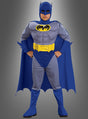 Ein Kind, das das „Kinderkostüm Batman Comicversion“ trägt – bestehend aus einem blauen Umhang, blauen Stiefeln, einem gelben Gürtel und einem grauen Anzug mit dem Batman-Logo auf der Brust, das an ein ikonisches DC-Comics-Lizenzkostüm angelehnt ist –, steht vor einem grauen Hintergrund.