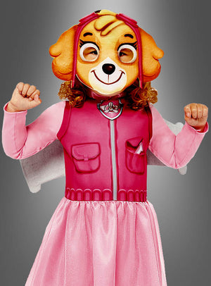Ein Kind posiert mit erhobenen Armen vor einem schlichten grauen Hintergrund. Es trägt das rosa „Skye“-Kostüm von Paw Patrol – komplett mit großer Hundemaske, rosa Weste und Rock – ein entzückendes Mädchenkostüm, das perfekt für jeden kleinen Skye-Fan ist.