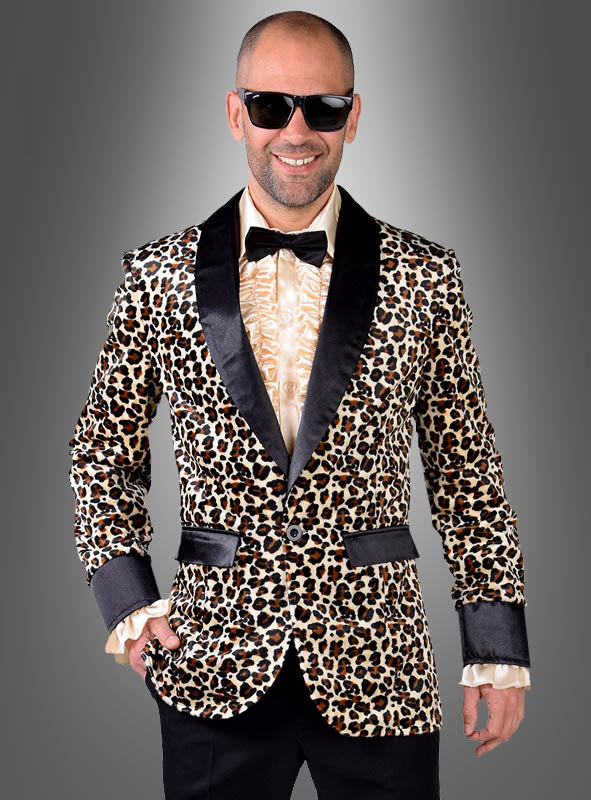 Ein Mann strahlt echte „Disco-Kostüm“-Atmosphäre aus, während er in einer auffälligen „Leoprint Jackett Deluxe“ – einem Herren-Sakko mit Leopardenmuster und eleganten schwarzen Revers – vor einem grauen Hintergrund mit Farbverlauf steht. Dazu trägt er ein cremefarbenes Hemd mit Rüschen, eine schwarze Hose, eine Fliege und eine Sonnenbrille.
