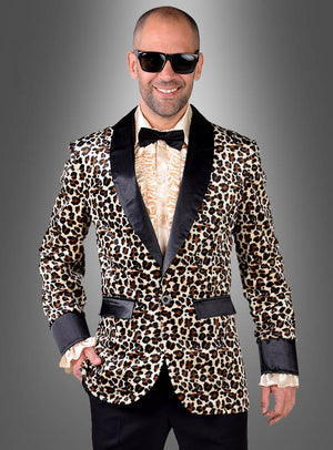 Ein Mann strahlt echte „Disco-Kostüm“-Atmosphäre aus, während er in einer auffälligen „Leoprint Jackett Deluxe“ – einem Herren-Sakko mit Leopardenmuster und eleganten schwarzen Revers – vor einem grauen Hintergrund mit Farbverlauf steht. Dazu trägt er ein cremefarbenes Hemd mit Rüschen, eine schwarze Hose, eine Fliege und eine Sonnenbrille.