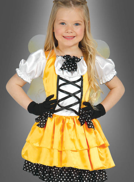 Ein junges Mädchen steht vor einem schlichten grauen Hintergrund, lächelt und trägt das Kostüm „Schmetterling gelb für Mädchen“, das sich durch ein gelb-schwarzes, von Schmetterlingen inspiriertes Design mit Rüschenrock, Puffärmeln, schwarzen Handschuhen und Flügeln auszeichnet.