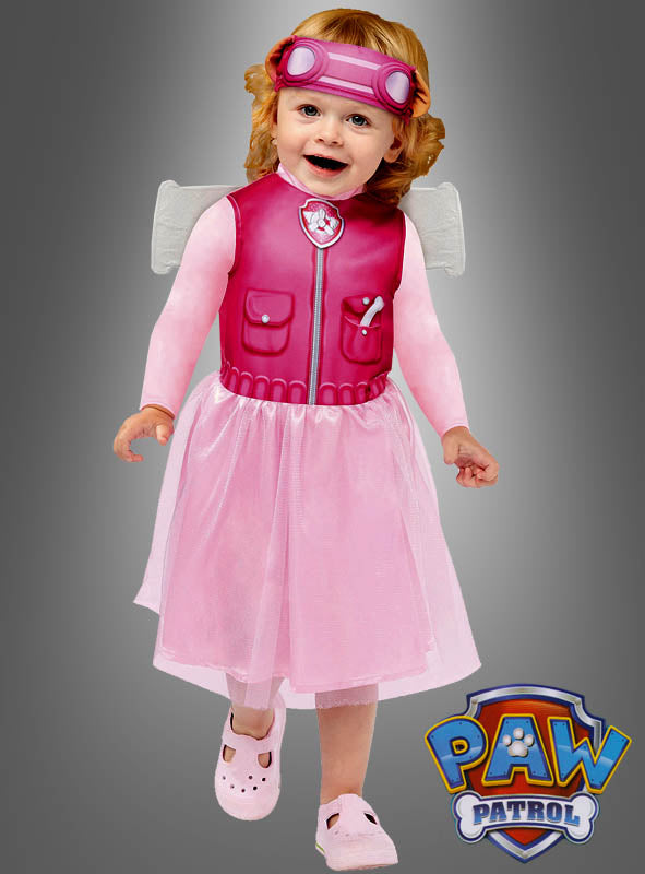 Ein kleines Kind im „Paw Patrol“-Skye-Kostüm, bestehend aus einer rosa Weste, einem Rock, einem Kopfschmuck, Flügeln und rosa Schuhen. Das „Paw Patrol“-Logo ist in der rechten unteren Ecke auf grauem Hintergrund zu sehen.