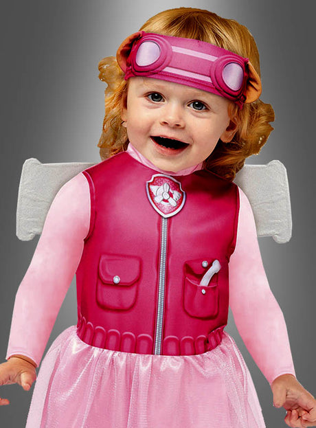 Ein kleines Kind trägt das „Paw Patrol“-Skye-Kostüm, komplett mit rosa Schutzbrille, einer Weste mit Pfotenabzeichen und glitzernden Feenflügeln – alles vor einem grauen Hintergrund. Damit ist es das perfekte Outfit für jeden kleinen Fan.