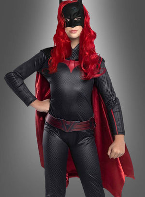 Eine Person, die das Original-Batwoman-Arrow-Kinderkostüm trägt – bestehend aus einem schwarz-roten Superheldenkostüm mit Maske, langer roter Perücke, Umhang und Handschuhen –, steht vor einem grauen Hintergrund.