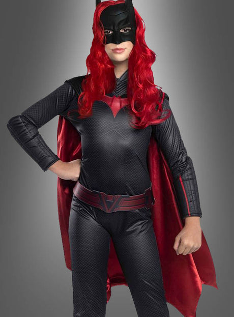 Eine Person, die das Original-Batwoman-Arrow-Kinderkostüm trägt – bestehend aus einem schwarz-roten Superheldenkostüm mit Maske, langer roter Perücke, Umhang und Handschuhen –, steht vor einem grauen Hintergrund.