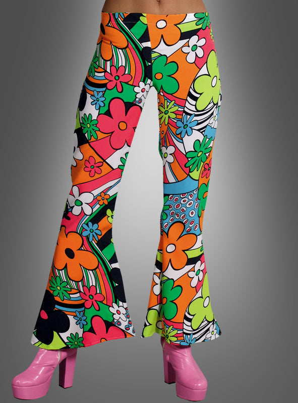 Eine Person, die die „Neon Flowers 70er Damen-Schlaghose“ mit einem bunten Blumenmuster trägt, kombiniert mit rosa Plateauschuhen, vor einem grauen Hintergrund.