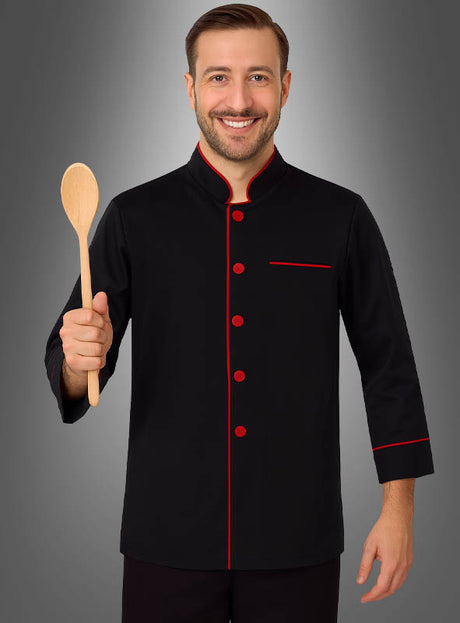 Ein Mann steht lächelnd vor einem grauen Hintergrund, hält einen Holzlöffel in der Hand und trägt die „Koch Jacke schwarz Unisex“ mit roten Zierstreifen.