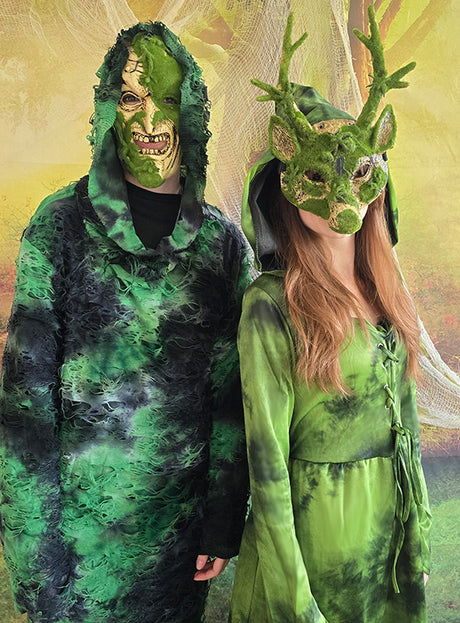 Zwei Personen in grünen Kostümen mit Waldmotiv stehen vor einem Waldhintergrund; eine trägt die „Mooshexe“-Version des Kapuzenkleids „Waldfee“ oder „Mooshexe“, komplett mit einer mit Moos verzierten Maske, während die andere das grüne Kapuzenkleid und eine Hirschmaske mit Geweih trägt.