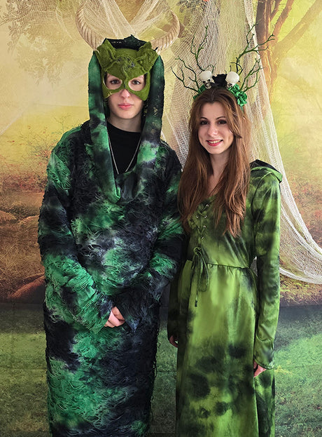 Zwei Personen in grünen Fantasiekostümen posieren vor einer Kulisse mit Waldmotiv; eine trägt eine Maske mit Hörnern und einen Umhang, während die andere das „Kapuzenkleid Waldfee oder Mooshexe“ mit einem geweihtartigen Kopfschmuck trägt.