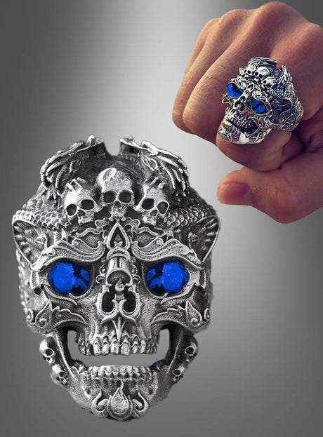 Der „Totenkopf Ring verstellbar silber“, ein silberner Ring in Totenkopfform mit auffälligen blauen Edelsteinaugen, wird sowohl an einer Faust als auch separat vor einem Hintergrund mit Farbverlauf präsentiert – und ist damit der perfekte Halloween-Schmuck.