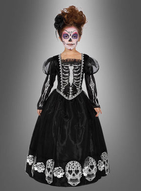 Ein Kind, das das „Deluxe Totenkopf-Kleid für Mädchen“ trägt – mit Skelettdetails und Totenköpfen am Saum – und eine von „Dia de los Muertos“ inspirierte Gesichtsbemalung trägt, steht vor einem grauen Hintergrund mit Farbverlauf.