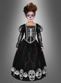 Ein Kind, das das „Deluxe Totenkopf-Kleid für Mädchen“ trägt – mit Skelettdetails und Totenköpfen am Saum – und eine von „Dia de los Muertos“ inspirierte Gesichtsbemalung trägt, steht vor einem grauen Hintergrund mit Farbverlauf.