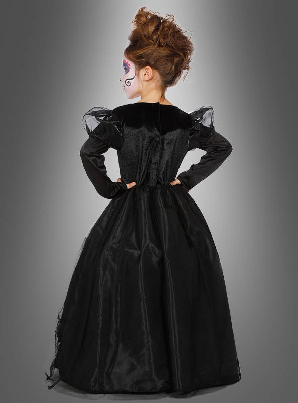 Ein Mädchen mit lockigem Haar und Gesichtsbemalung steht mit den Händen in den Hüften, den Rücken zur Kamera gewandt, und trägt das „Deluxe Totenkopf Kleid für Mädchen“ – ein langes schwarzes Skelettkleid – vor einem schlichten grauen Hintergrund.