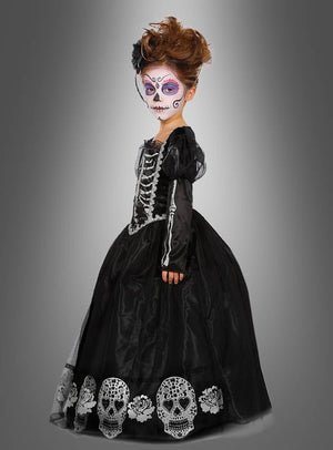 Ein Kind trägt das „Deluxe Totenkopf-Kleid für Mädchen“, ein langes schwarzes Kleid mit Totenkopf-Motiven, sowie eine Gesichtsbemalung im Sugar-Skull-Stil und steht vor einem grauen Hintergrund mit Farbverlauf.