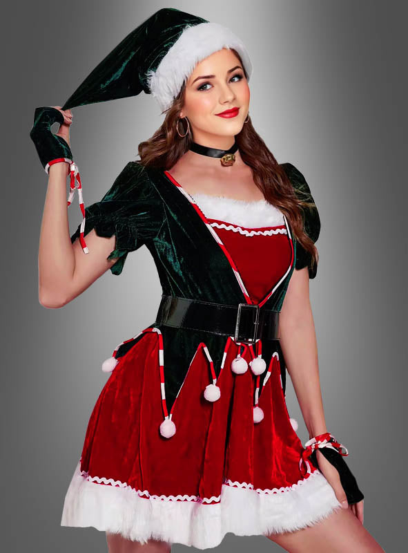 Eine Frau, die ein sexy Weihnachtself-Kostüm für Damen trägt – bestehend aus einem vom Weihnachtsmann inspirierten Kleid und einer Mütze sowie schwarzen Handschuhen und einem Gürtel –, posiert vor einem schlichten grauen Hintergrund.