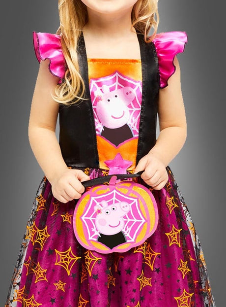 Ein kleines Kind, das ein Peppa-Pig-Hexe-Kostüm mit Spinnennetzmustern trägt und eine dazu passende, kürbisförmige Tasche in der Hand hält.